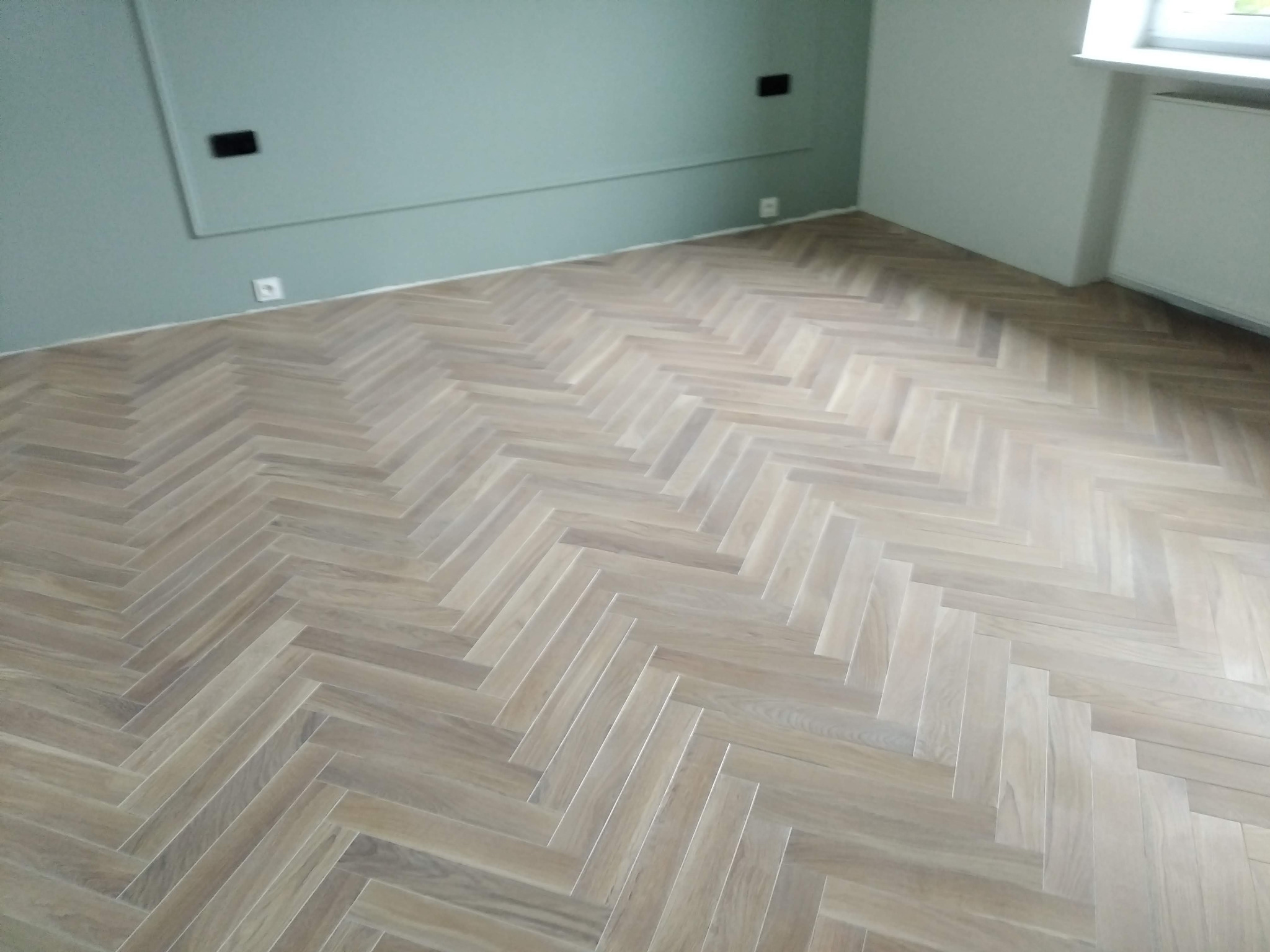 Parkiet ułożony w jodełkę na podłodze w pokoju z zielonymi i białymi ścianami, widoczne gniazdka elektryczne i grzejnik pod oknem.