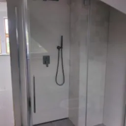 Nowoczesna kabina prysznicowa z deszczownicą i szarymi płytkami w łazience na poddaszu, widoczne szklane drzwi i szara podłoga imitująca drewno.