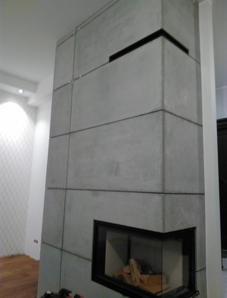 Nowoczesny kominek obłożony szarymi płytami imitującymi beton, z czarnym obramowaniem paleniska i wkładem kominkowym, widoczny fragment podłogi i ściany z tapetą w tle.