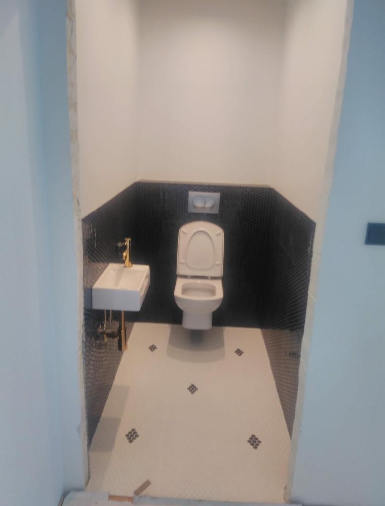 Nowoczesna toaleta w trakcie wykańczania: biała umywalka z złotą armaturą, wisząca muszla klozetowa, czarne mozaikowe płytki na ścianie i biała podłoga z dekoracyjnymi wstawkami.