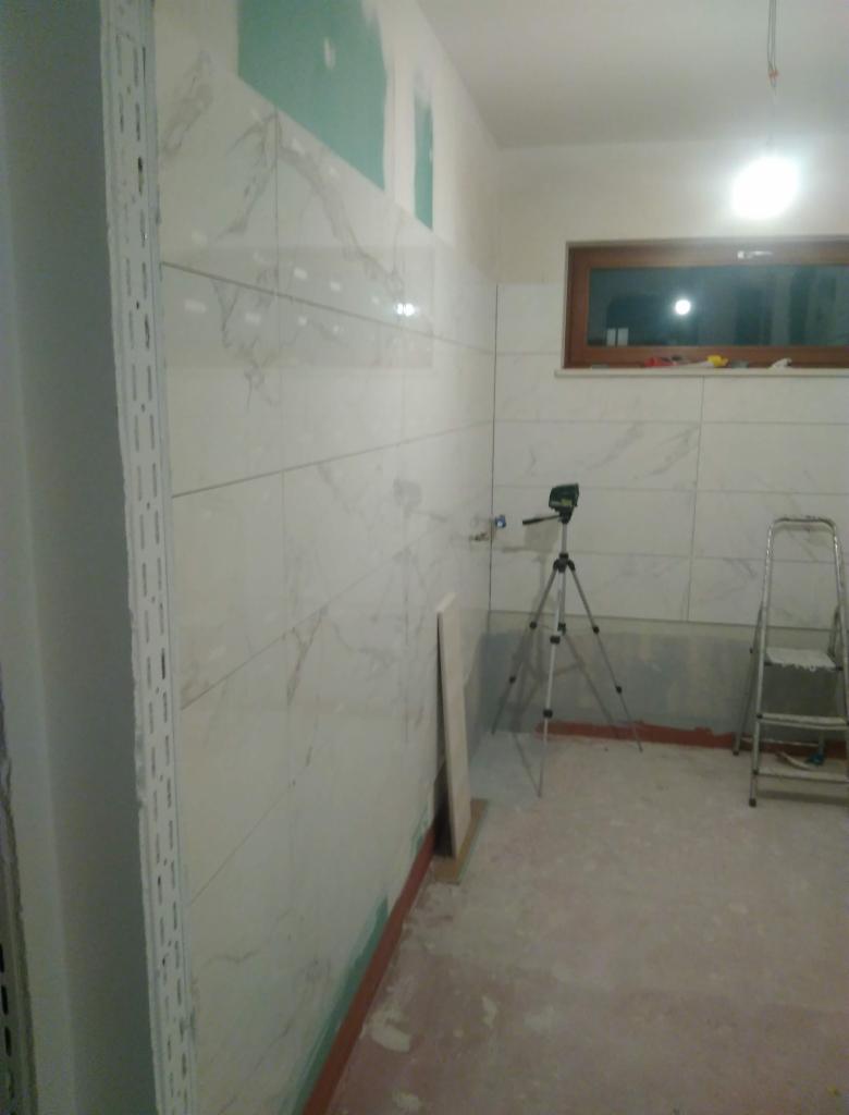 Wnętrze łazienki w trakcie układania białych, marmurkowych płytek ściennych, widoczny laser krzyżowy na statywie, drabina i fragment płyty kartonowo-gipsowej przy podłodze.