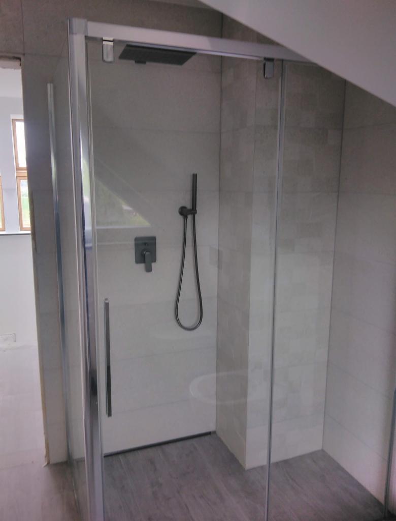 Nowoczesna kabina prysznicowa z deszczownicą i szarymi płytkami w łazience na poddaszu, widoczne szklane drzwi i szara podłoga imitująca drewno.