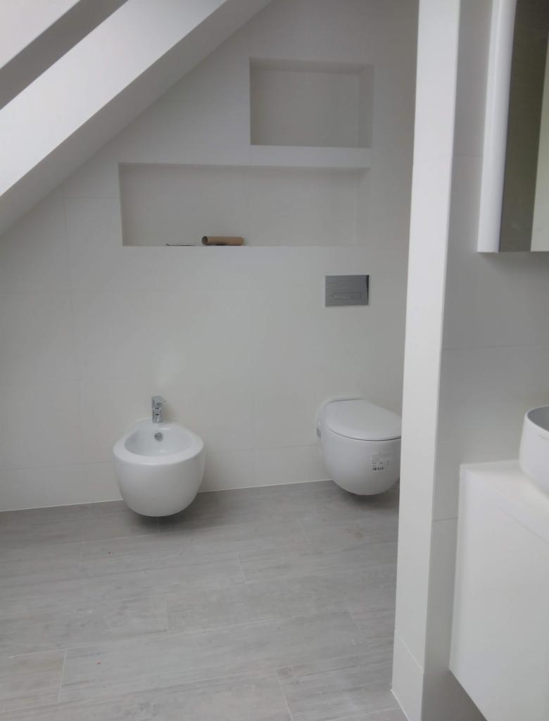 Nowoczesna, minimalistyczna łazienka na poddaszu z białymi płytkami, wiszącą miską WC i bidetem, oraz wnękami w ścianie, jedna z rolką po papierze toaletowym.