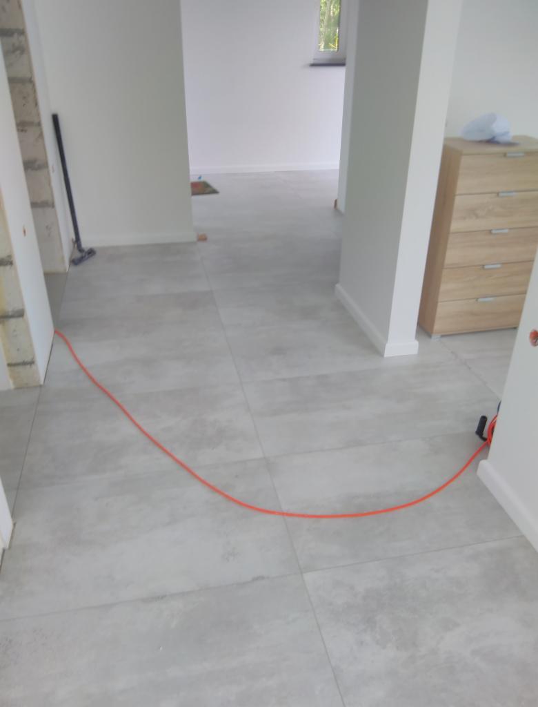 Jasnoszare płytki podłogowe ułożone w nowym domu, widoczny kabel elektryczny na podłodze, w tle meble i otwarte przejście do innego pomieszczenia.