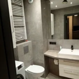 Nowoczesna łazienka w szarościach z podwieszaną toaletą, pralką, grzejnikiem drabinkowym i umywalką z szafką, oświetlona punktowymi lampami i czarnym reflektorem.