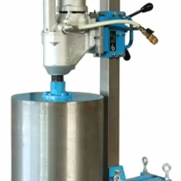 Stacjonarna wiertnica diamentowa z cylindrycznym wiertłem koronowym, zamocowana na niebieskim, metalowym stelażu, widoczne przewody i dźwignia sterująca.