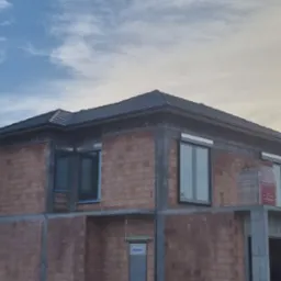 Surowy stan domu z czerwonej cegły ceramicznej, widoczne okna z roletami zewnętrznymi, dach pokryty dachówką ceramiczną, brak ocieplenia, na drugim planie błękitne niebo z chmurami.