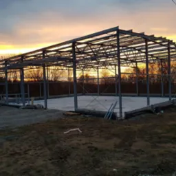 Szkielet hali z belek stalowych na betonowej podstawie, widok z zewnątrz o zmierzchu, pomarańczowe niebo w tle.