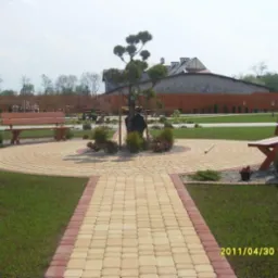 Ogród z alejką z kostki brukowej w odcieniach beżu i czerwieni, prowadzącą do okrągłego placu z drzewem bonsai i rzeźbą, otoczony zielonym trawnikiem, ławkami i placem zabaw w tle.