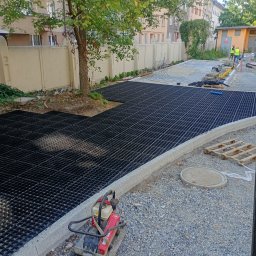 RT BRUK Kompleksowe Usługi Brukarskie - Montaż ażurowych płyt na podbudowie z kruszywa, obrzeża z kostki granitowej. W tle budynki mieszkalne i pracownicy w odblaskowych kamizelkach.