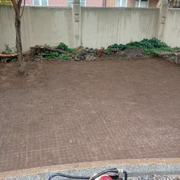 RT BRUK Kompleksowe Usługi Brukarskie - Przygotowany teren pod kostkę brukową z ubijarką w rogu. Widoczne ślady po kostce, betonowy mur w tle i resztki materiałów budowlanych.