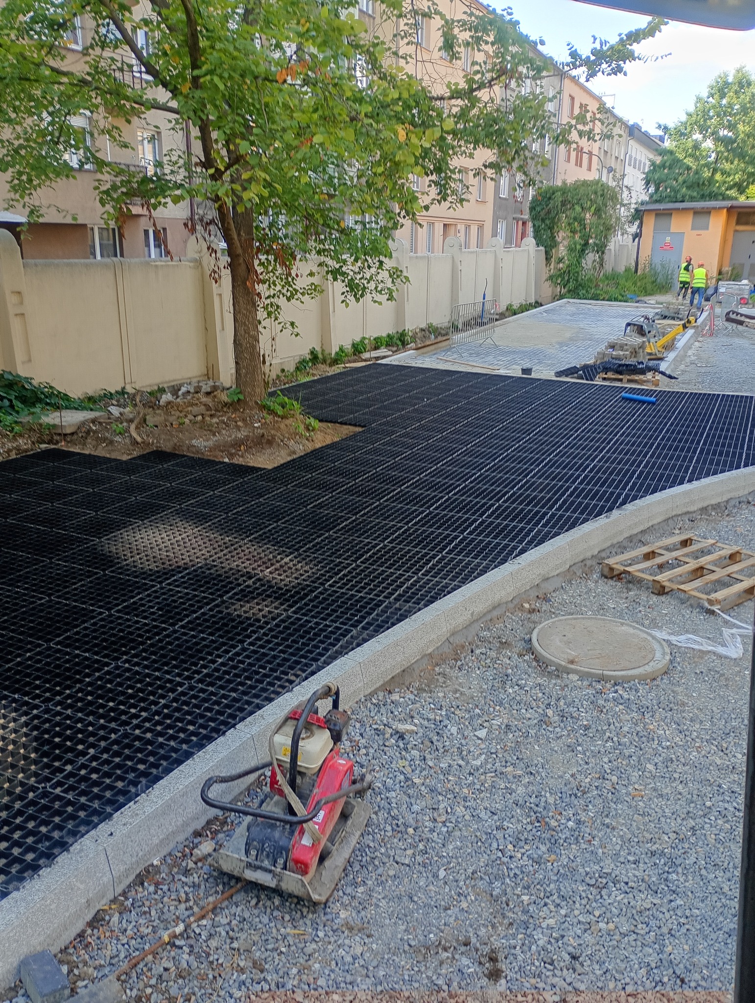 Montaż ażurowych płyt na podbudowie z kruszywa, obrzeża z kostki granitowej. W tle budynki mieszkalne i pracownicy w odblaskowych kamizelkach.