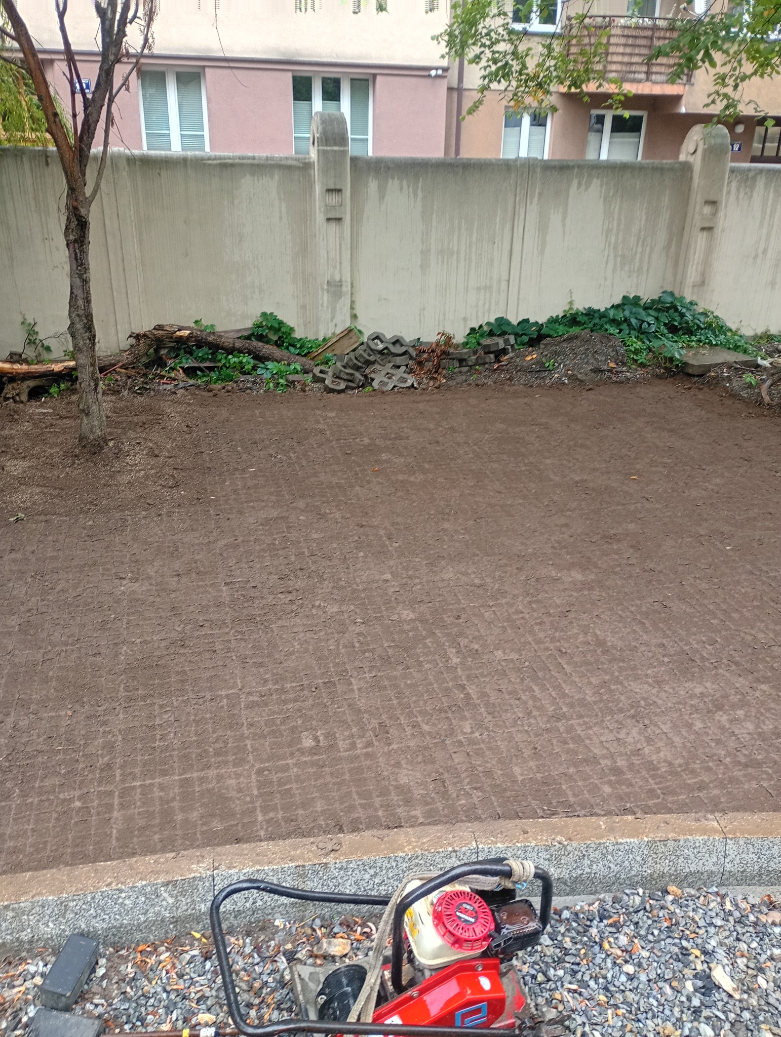 Przygotowany teren pod kostkę brukową z ubijarką w rogu. Widoczne ślady po kostce, betonowy mur w tle i resztki materiałów budowlanych.