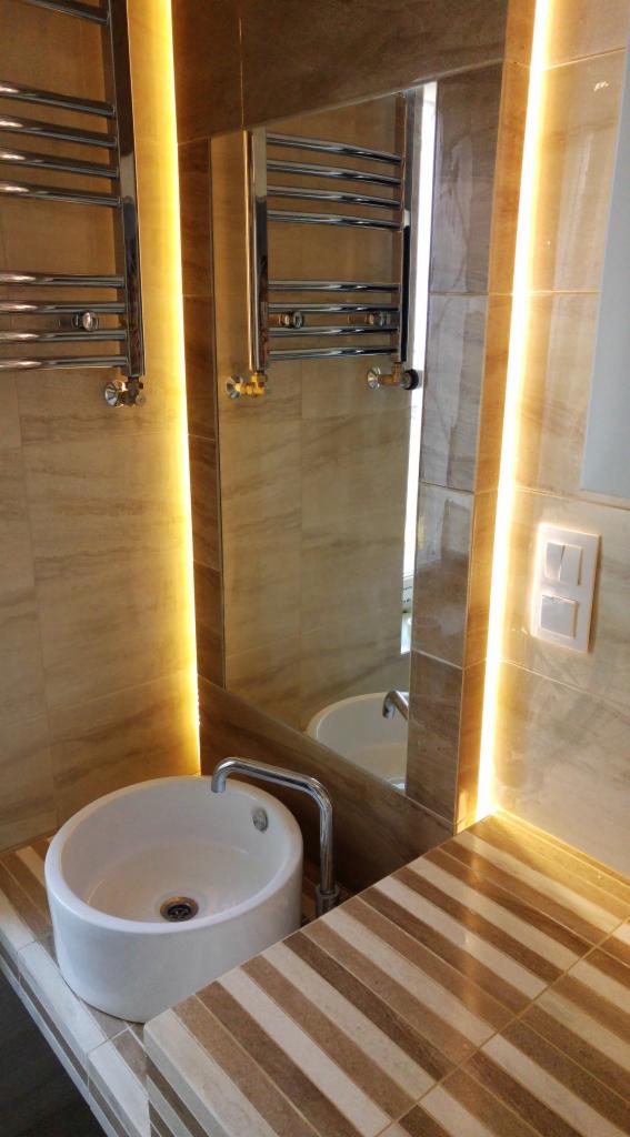 Nowoczesna łazienka z okrągłą umywalką nablatową, podświetlanym lustrem i grzejnikiem drabinkowym, wykończona beżowymi płytkami imitującymi marmur i dekoracyjnymi płytkami w paski na blacie.