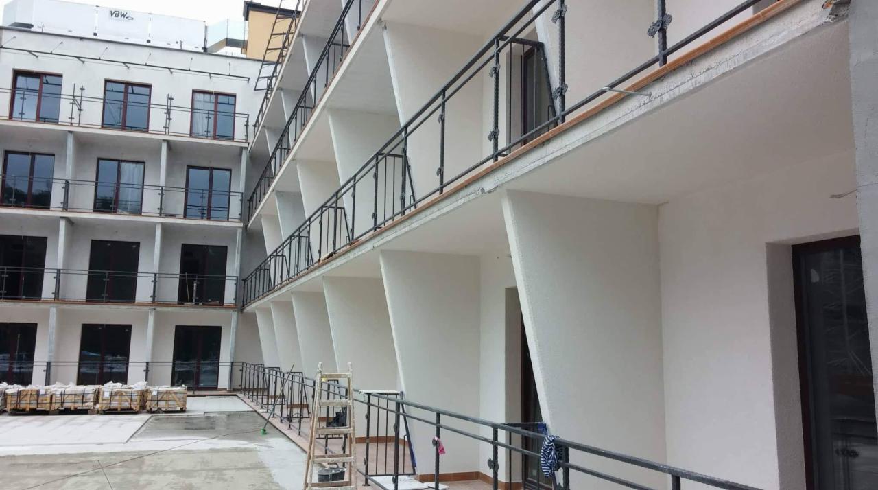 Budynek wielorodzinny z balkonami w trakcie wykończenia, widoczne rusztowania i materiały budowlane, elewacja w jasnym kolorze.