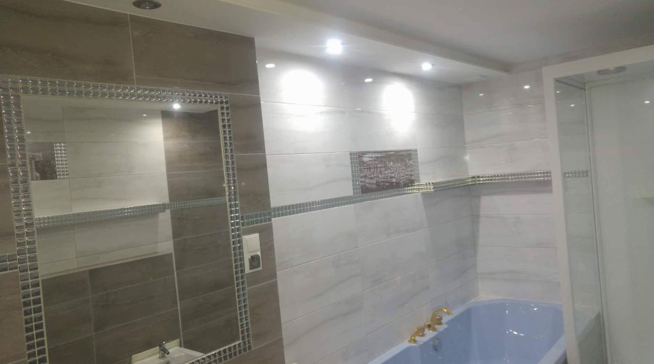 Nowoczesna łazienka z beżowo-białymi płytkami, lustrem w ozdobnej ramie i niebieską wanną z pozłacaną armaturą. Dekoracyjny pasek z mozaiki oddziela poziomy płytek. Na ścianie wisi obrazek.