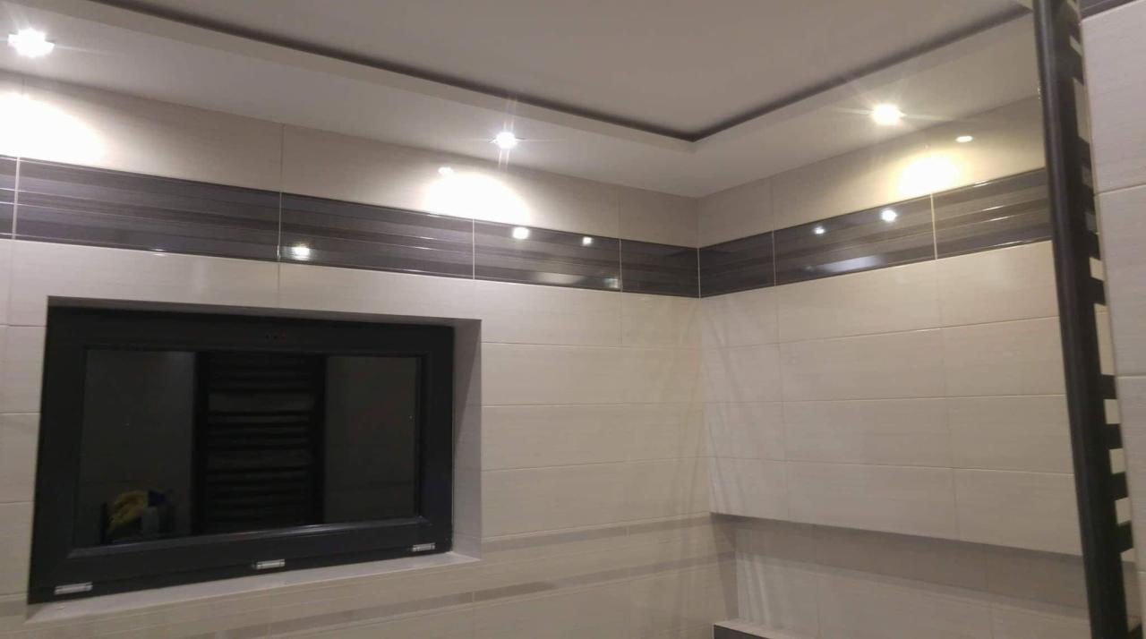 Wykończona łazienka z beżowymi płytkami, ciemnoszarym pasem dekoracyjnym i czarną ramą okienną, oświetlona punktowymi lampami sufitowymi.