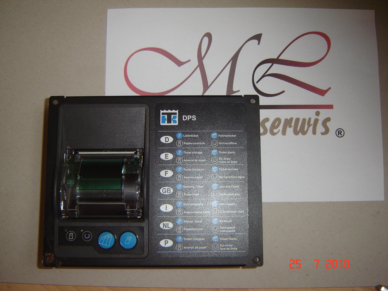 Czarny panel sterowania urządzenia DPS z widocznym mechanizmem drukującym rolkę papieru, na tle kartki z logo serwisu z datą 25 7 2010.
