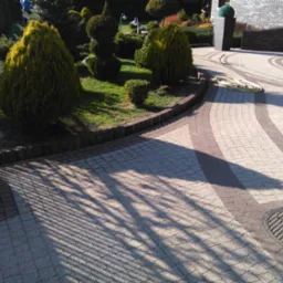 Aranżacja posesji z ścieżką z kostki w odcieniach szarości i brązu, obrzeża z kostki, trawnik z ozdobnymi krzewami i drzewami. Wąż ogrodowy leży zwinięty obok ścieżki.