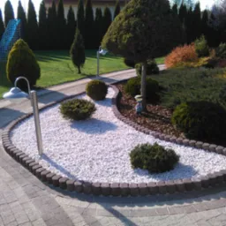 Ogród z ścieżką z jasnej kostki, obrzeża z ciemnej kostki, lampy ogrodowe, ozdobne krzewy i trawnik. W tle wysokie drzewa iglaste.