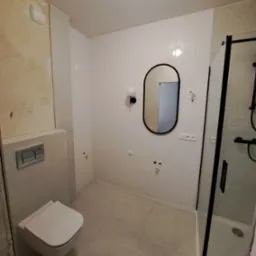 Biała łazienka z owalnym lustrem w czarnej ramie, kabiną prysznicową z czarnymi elementami i podwieszaną toaletą przed wykończeniem ściany.