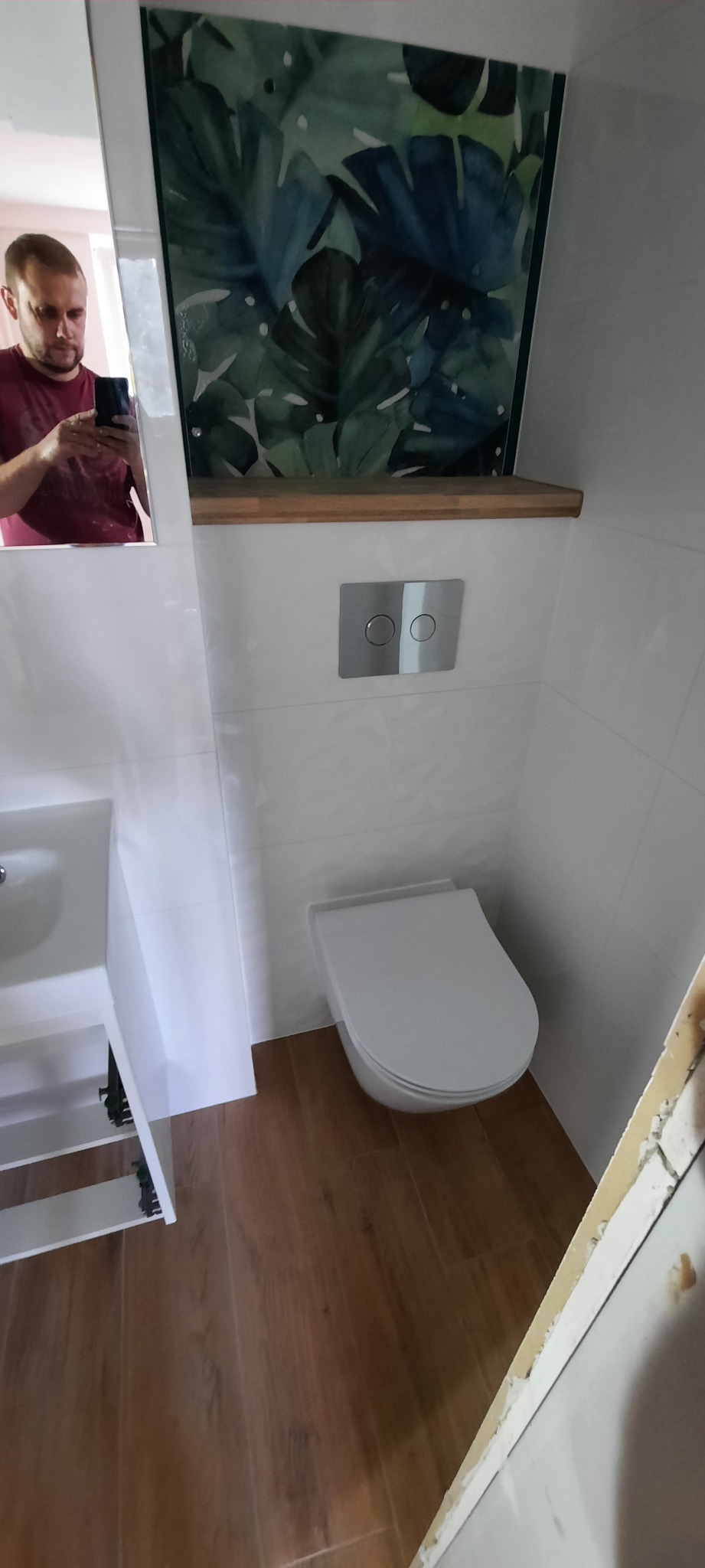 Nowoczesna łazienka po remoncie: białe płytki, drewnopodobna podłoga, wisząca toaleta z chromowanym przyciskiem spłukiwania, półka z drewnianym blatem i obraz z motywem roślinnym. W lustrze odbicie...