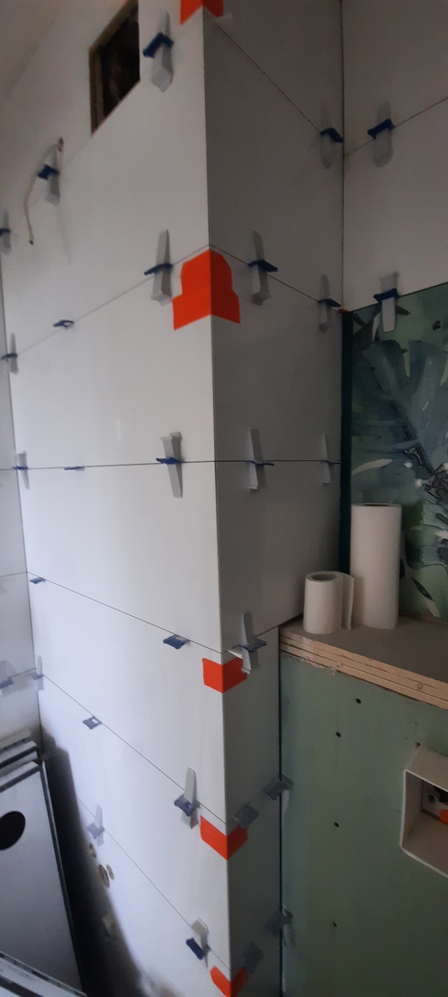 Białe płytki ścienne w trakcie układania, z wykorzystaniem krzyżyków dystansowych i pomarańczowej taśmy na narożnikach, widoczne rolki papieru i płyta gipsowo-kartonowa