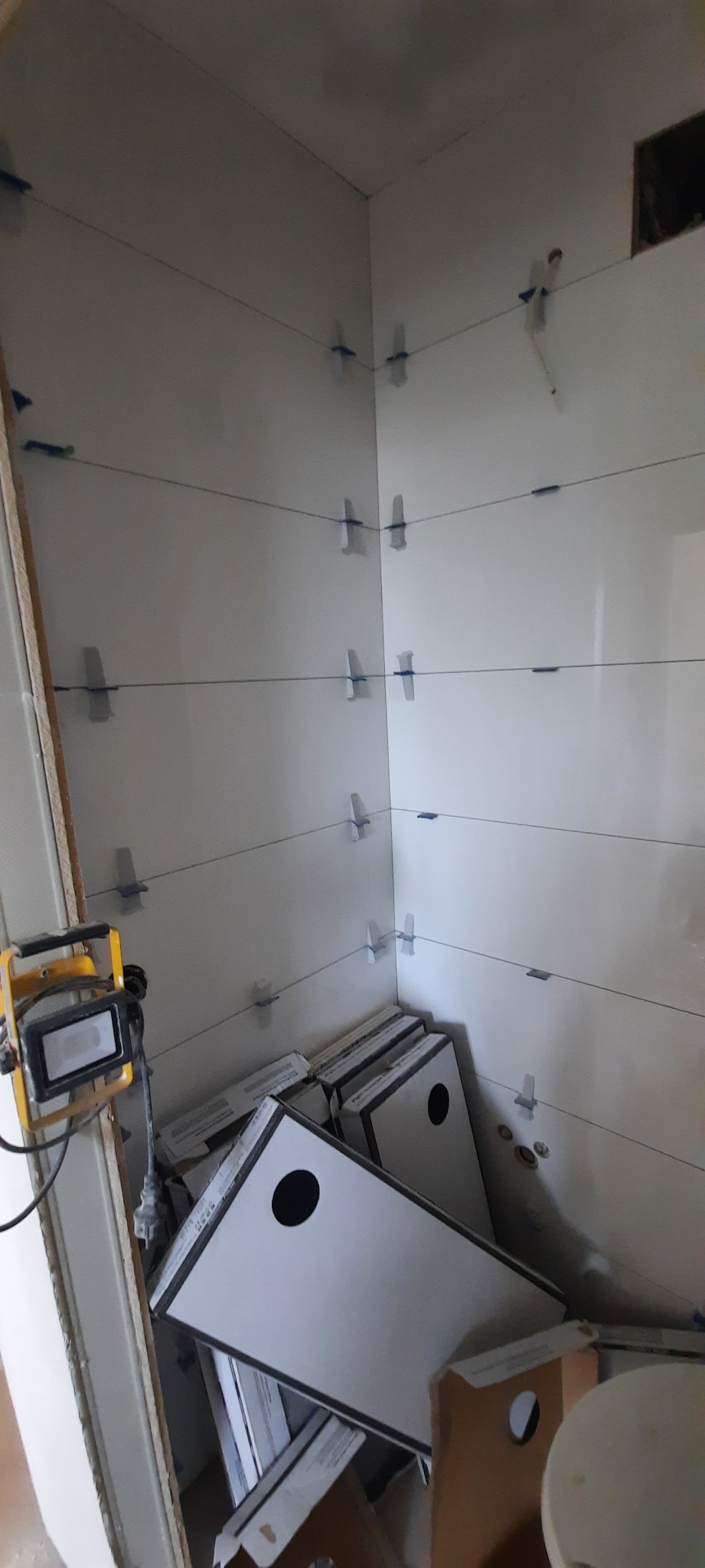 Białe płytki ścienne w trakcie układania w rogu pomieszczenia, widoczne krzyżyki dystansowe, poziomice laserowe i kartony z płytkami.