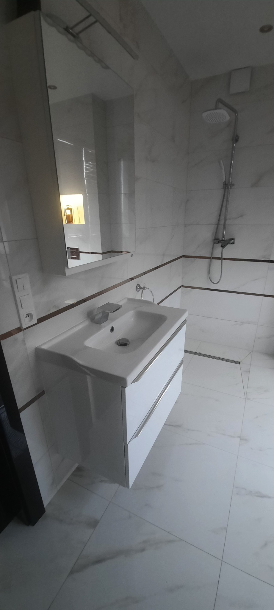 Nowoczesna łazienka z białymi marmurkowymi płytkami, umywalką z szafką w kolorze białym, lustrem z oświetleniem odbijającym ciepłe światło, kabiną prysznicową z deszczownicą i odpływem liniowym...