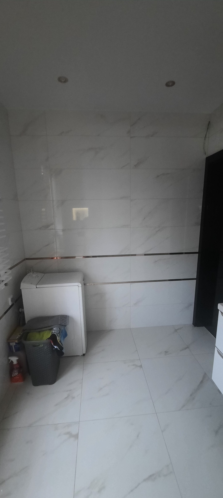 Ukończona łazienka z białymi marmurkowymi płytkami na ścianach i podłodze, widoczna pralka, kosz na pranie i fragment szafki