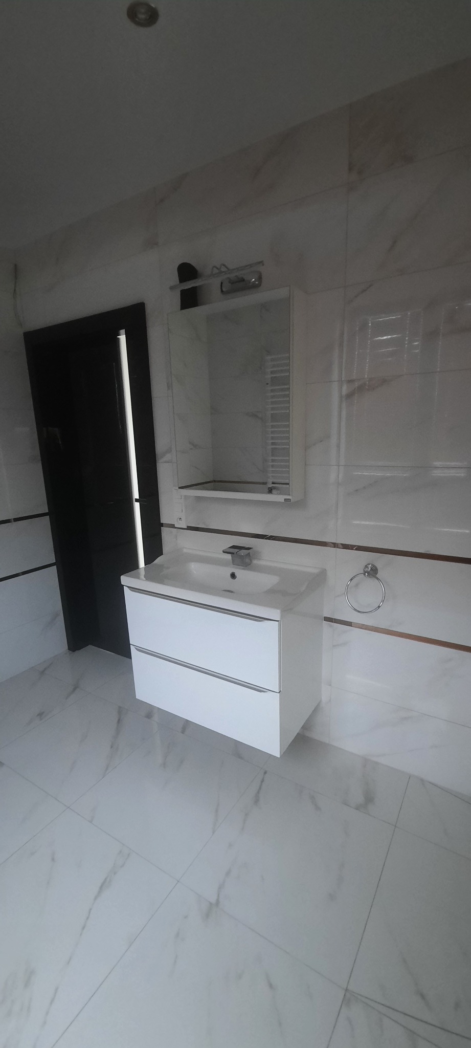 Nowoczesna łazienka po remoncie: białe marmurowe płytki, biała szafka pod umywalkę z dwiema szufladami, lustro z oświetleniem i czarne drzwi.