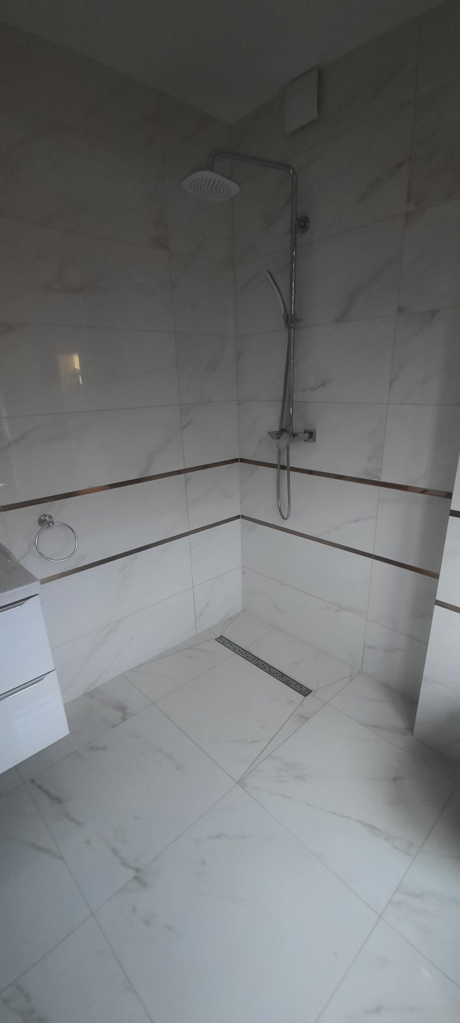 Nowoczesna kabina prysznicowa z białymi płytkami imitującymi marmur, ozdobnymi złotymi paskami, deszczownicą i odpływem liniowym.