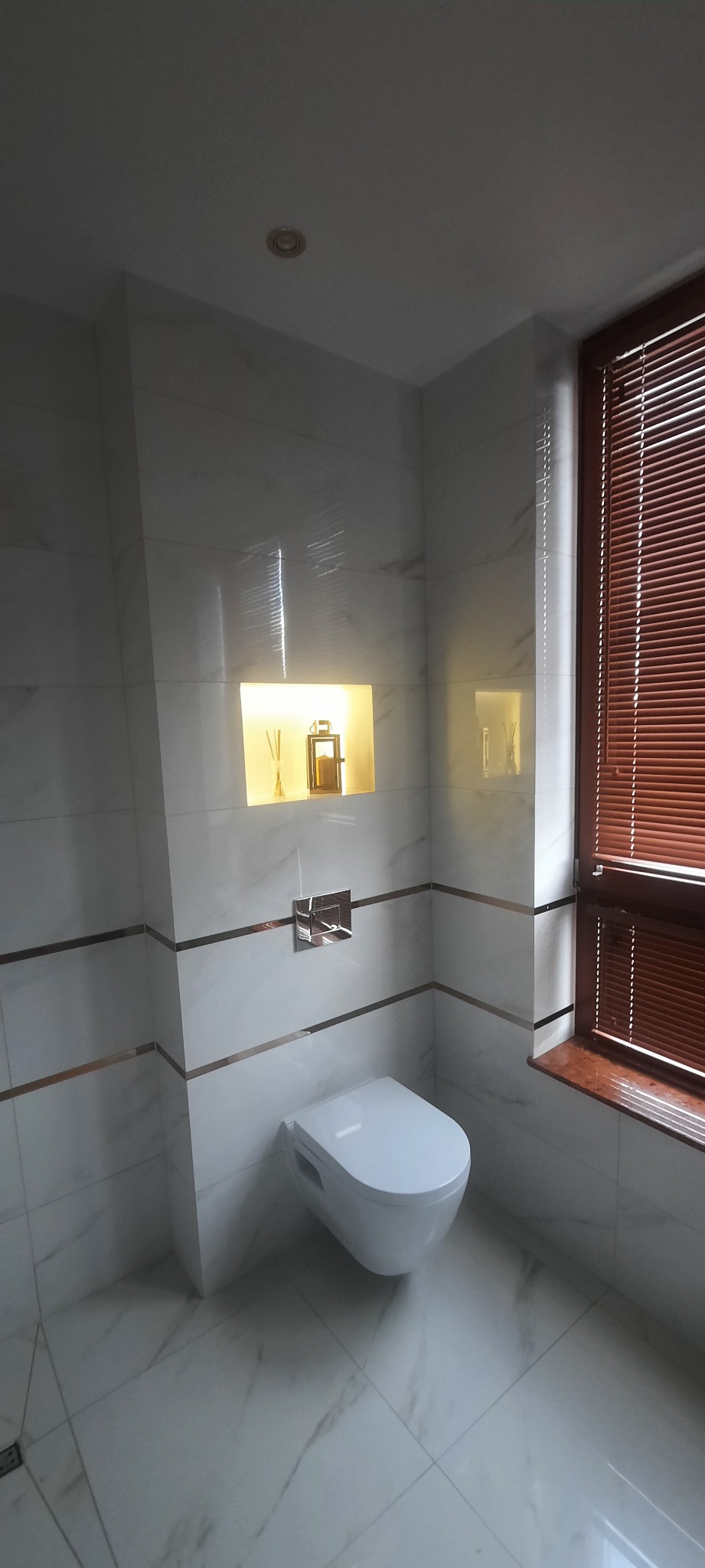 Nowoczesna łazienka z białymi marmurkowymi płytkami, podwieszaną toaletą i podświetlaną niszą z dekoracjami, okno z drewnianymi żaluzjami.