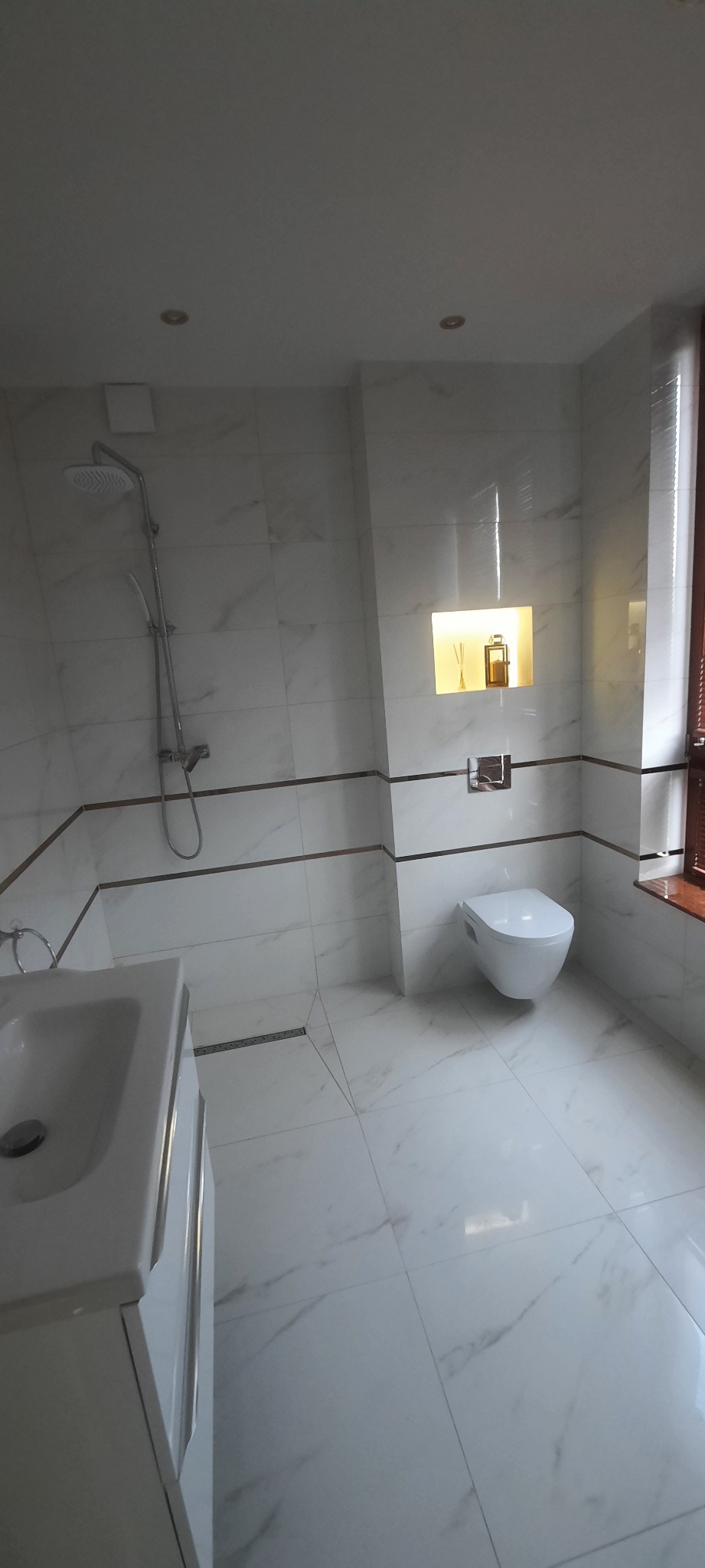 Elegancka łazienka z białymi marmurkowymi płytkami, kabiną prysznicową typu walk-in, wiszącą toaletą i wnęką z dekoracyjnymi akcesoriami.