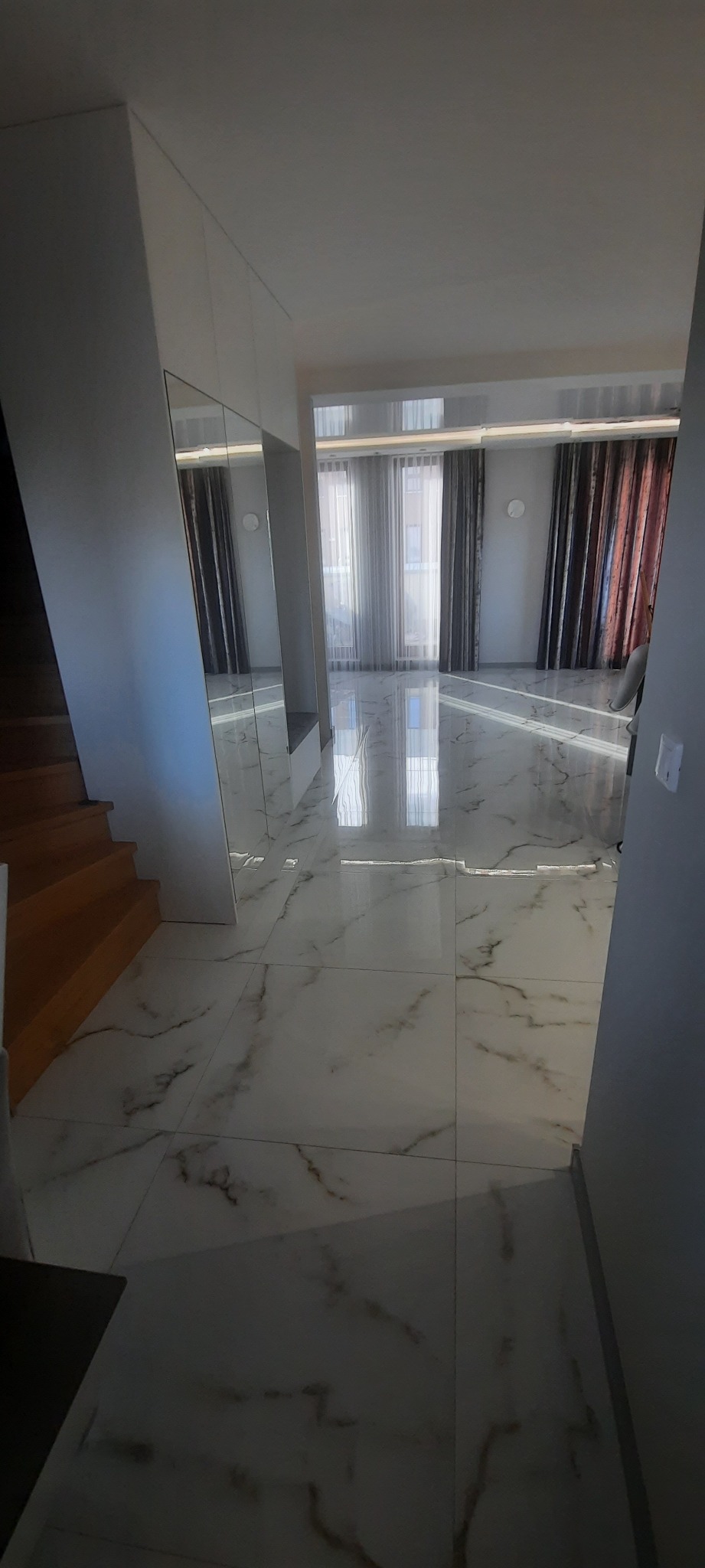 Nowoczesny korytarz z marmurową posadzką w odcieniach bieli i szarości, lustrzana szafa w zabudowie, widok na salon z zasłonami w dwóch kolorach, drewniane schody po lewej stronie.
