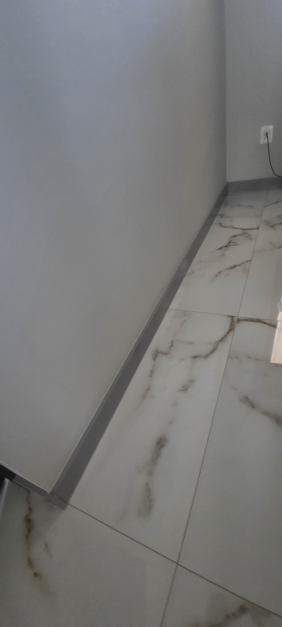 Ułożone płytki podłogowe imitujące marmur z szarą listwą przypodłogową i gładką, szarą ścianą z gniazdkiem elektrycznym.