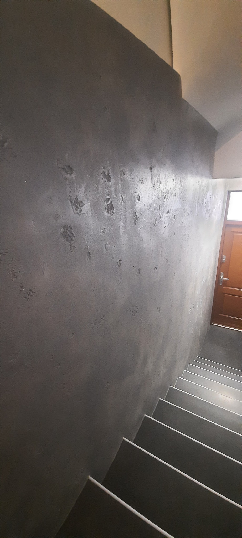 Ściana klatki schodowej z dekoracyjną szarą fakturą, widoczne schody i fragment drzwi wejściowych.