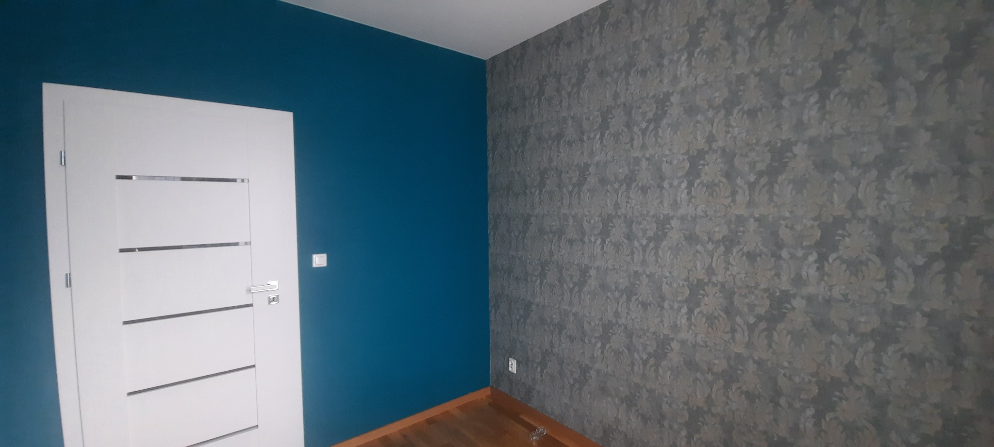 Pokój po remoncie: ściana pomalowana na intensywny niebieski kolor, białe drzwi z poziomymi, srebrnymi wstawkami oraz ściana pokryta tapetą w kwiatowy wzór w odcieniach szarości.