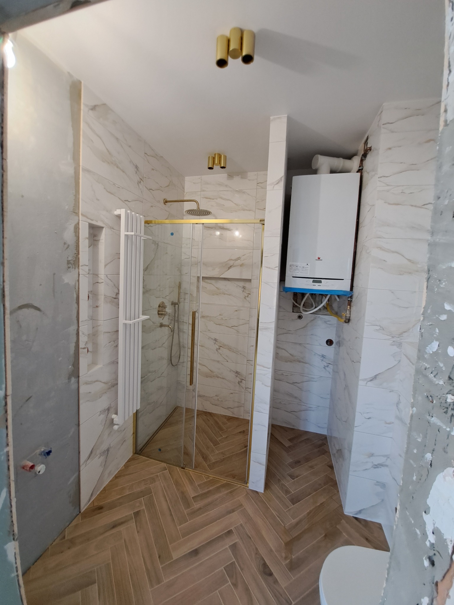 Wnętrze łazienki w trakcie prac wykończeniowych, widoczne marmurowe płytki, kabina prysznicowa ze złotymi detalami, grzejnik drabinkowy oraz piec gazowy z niebieskim zabezpieczeniem.