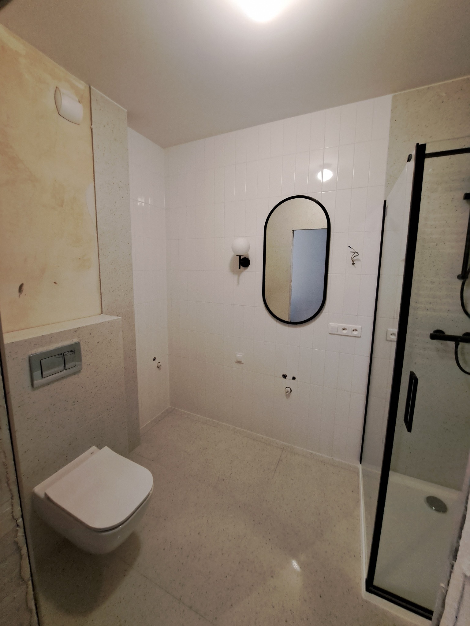 Biała łazienka z owalnym lustrem w czarnej ramie, kabiną prysznicową z czarnymi elementami i podwieszaną toaletą przed wykończeniem ściany.