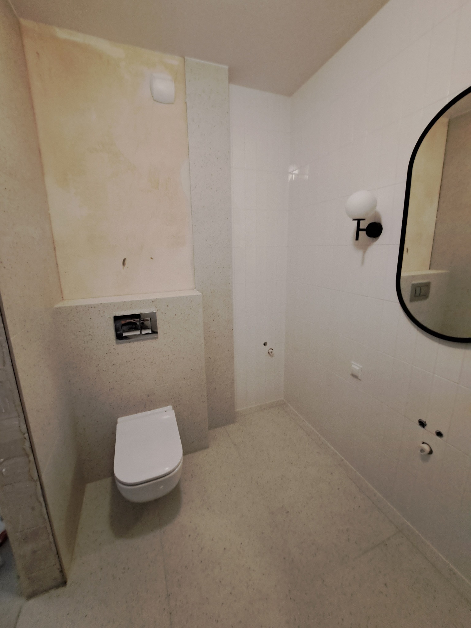 Niedokończona łazienka z podtynkową toaletą, surową ścianą przygotowaną pod wykończenie, białą glazurą i okrągłym lustrem z designerską lampą.