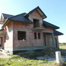 Surowy stan domu jednorodzinnego z czerwonej cegły z czarnym dachem, widoczne niezamknięte otwory okienne i drzwiowe, na pierwszym planie trawa i skrzynka elektryczna.