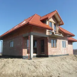 Surowy, dwukondygnacyjny dom z czerwonej cegły i czerwoną dachówką, z niezadaszonym balkonem i betonowym podcieniem wejściowym, w trakcie budowy, na nierównym terenie z widocznym wykopem.