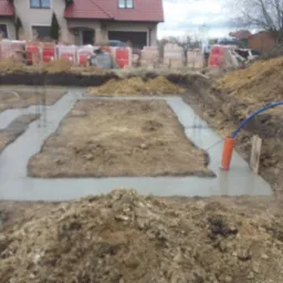 Wylane fundamenty pod budynek jednorodzinny, widoczne zbrojenia i przygotowane materiały budowlane w tle, w tym cegły i pustaki.