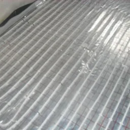 Białe rurki ogrzewania podłogowego ułożone równolegle na folii z czerwoną siatką, widoczne mocowania rurek.