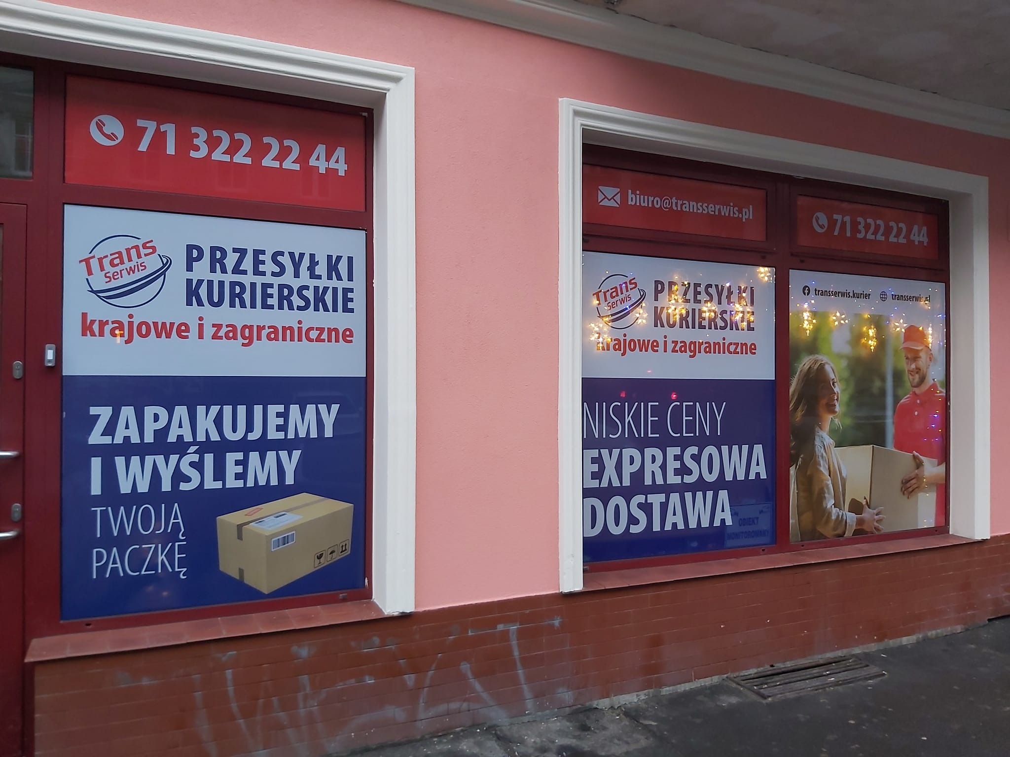 Fasada budynku z dwoma oknami oklejonymi reklamami firmy kurierskiej, oferującej przesyłki krajowe i zagraniczne, z widocznymi numerami telefonów i adresem e-mail.