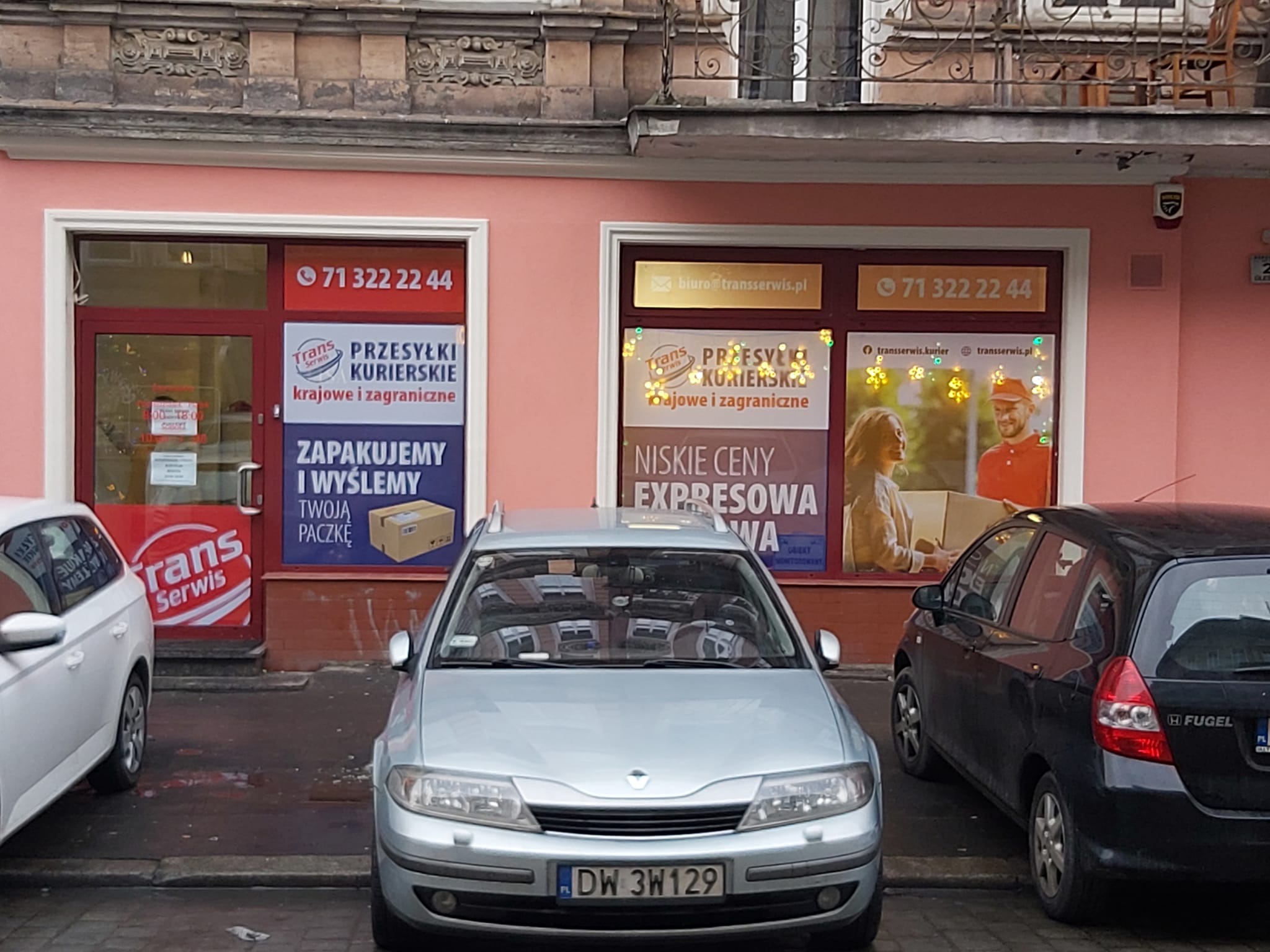 Fasada punktu kurierskiego Trans Serwis we Wrocławiu z reklamami przesyłek krajowych i zagranicznych, zaparkowane samochody przed wejściem.