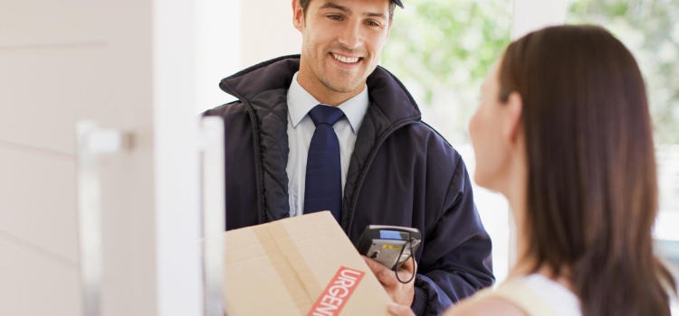 Uśmiechnięty kurier w czapce i granatowej kurtce wręcza kartonowe pudełko z napisem 'URGENT' klientce, używając terminala płatniczego.