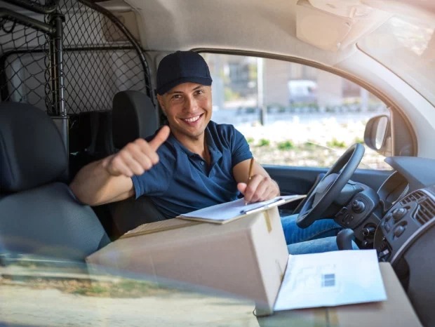 Uśmiechnięty kierowca w kabinie dostawczego samochodu, z uniesionym kciukiem, trzymający długopis i notatnik na paczce, prawdopodobnie po zakończonym kursie.