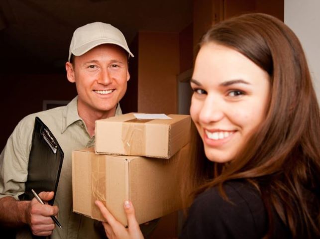 Uśmiechnięty kurier w czapce i koszuli trzymający czarny notatnik z długopisem przekazuje dwa kartonowe pudełka uśmiechniętej kobiecie w drzwiach.